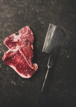 Flat-lay of fresh uncooked beef meat t-bone steaks and butchers cleaver over rough black stone background, top view. Butchers shop concept